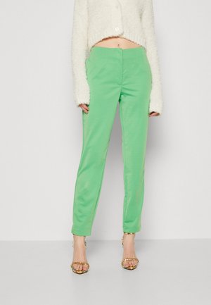 Vrouw met hoge taille lichtgroene broek, een pluche cropped witte trui en open-toe heels met gouden kettingbandjes, staand tegen een witte achtergrond.