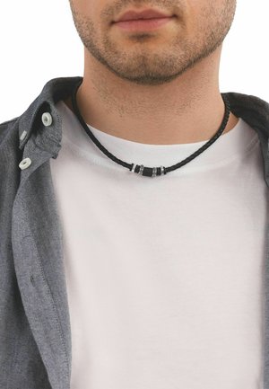 Close-up van een man die een zwart gevlochten ketting met metalen kralen draagt, een wit T-shirt en een open grijs overhemd met knopen.
