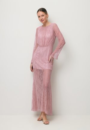 Vrouw die een lange doorschijnende roze kanten jurk met lange mouwen en naaktkleurige sandalen met hakken draagt, staand tegen een effen witte muur.