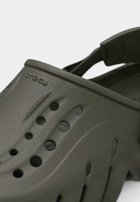 Gros plan de Crocs vert olive avec des trous de ventilation et une bride de talon réglable sur un fond uni.
