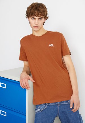T-shirt en coton à manches courtes de couleur rouille, avec un logo blanc sur la poitrine gauche. Porté avec un jean en denim bleu.