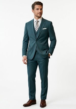 Hombre con traje tres piezas color verde azulado, camisa blanca, corbata estampada, pañuelo blanco en el bolsillo y zapatos de vestir marrones, de pie con una mano en el bolsillo.