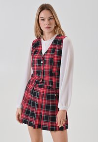 Completo a vest e gonna in tartan rosso e nero con una vestibilità sartoriale; presenta una chiusura con bottoni e un design plissettato. Lo sfondo è bianco semplice.