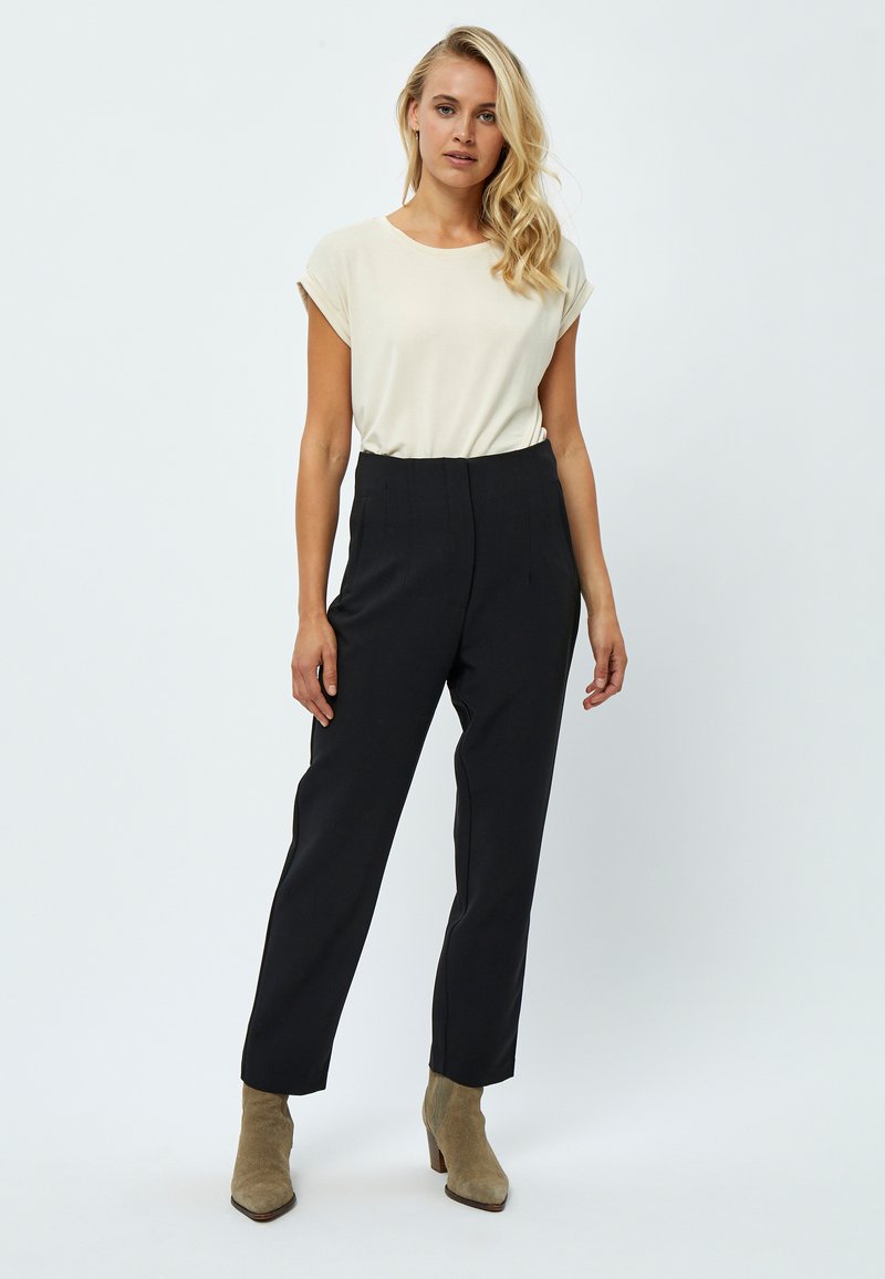 Top beige a maniche corte abbinato a pantaloni neri a vita alta e stivaletti. I pantaloni presentano dettagli plissettati e una vestibilità sartoriale.