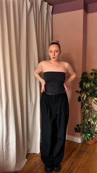 Femme aux cheveux attachés en chignon, portant un haut noir sans bretelles et un pantalon large noir, debout les mains sur les hanches dans une pièce aux murs roses avec une plante.