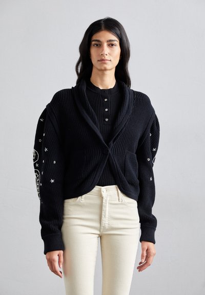 Cardigan nero lavorato a maglia con colletto a scialle, chiusura con bottoni e maniche lunghe, decorato con ricami bianchi a forma di stella, abbinato a jeans chiari.
