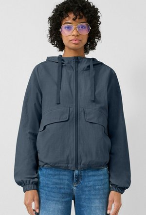 Frau mit lockigem Haar trägt eine blaue Kapuzenjacke mit Reißverschluss, blaue Jeans und große, lila getönte Brille, blickt nach vorne vor einem schlichten Hintergrund.