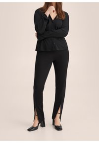 Camisa negra de botones con mangas largas, combinada con pantalones negros a medida con aberturas laterales y zapatos negros de tacón bloque. Textura suave.
