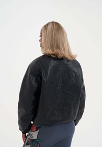 Černá kožená bunda s vyraženým vzorem draka na zadní straně, s volným střihem a minimálním množstvím kovových doplňků. Dlouhé rukávy a strukturovaný povrch.