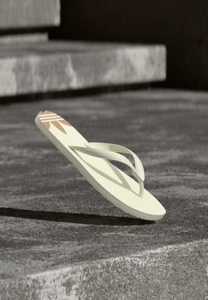 Enkele beige teenslipper met bandje en geometrisch ontwerp die rust op betonnen trappen in zonlicht, en werpt een schaduw.