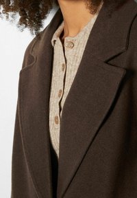 Primo piano di un cappotto di lana marrone sopra un cardigan a coste beige con bottoni beige su una persona con capelli ricci.