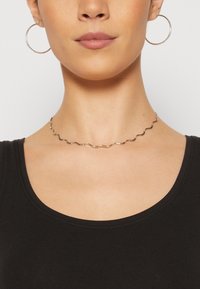 Srebrna valovita choker ogrlica ističe metalni sjaj. Nosena s velikim okruglim naušnicama protiv crnog majice. Glatka tekstura.