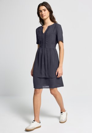 Robe bleu marine à manches courtes avec un décolleté en V, détail plissé et jupe superposée. Associe avec des baskets blanches, présentant un style décontracté.