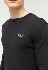 Mann trägt einen schwarzen Rundhalsausschnitt-Pullover mit kleinem weißen eingesticktem "SuperDry"-Logo und asiatischen Zeichen auf der linken Brust.