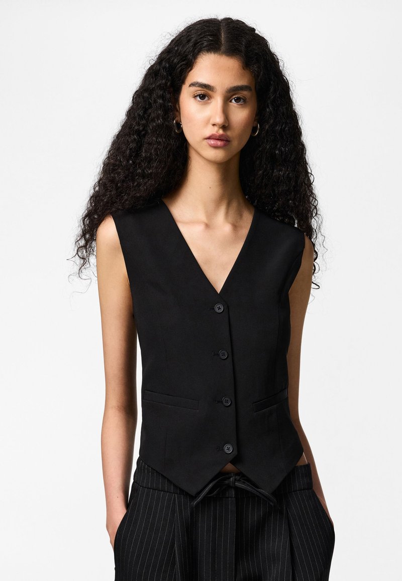 Jauna sieviete ar gariem cirtainiem matiem valkā melnu bezroku pogas vesti un melnas svītrainas bikses, rokas kabatās.