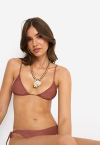 Τρίγωνο μπικίνι με πλεξούδα σε μεταλλικό ροζ χρώμα, με λεπτές τιράντες και δεμένο στη μέση. Διακοσμημένο με ένα χοντρό κολιέ με κοχύλια και χρυσούς τόνους.