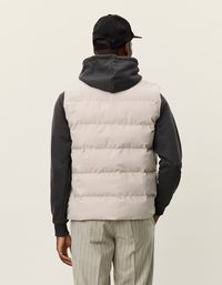 Πανωφόρι σε μπεζ χρώμα με επένδυση, πάνω από σκουρόχρωμο γκρι φούτερ, με λεία υφή και οριζόντιες επενδυμένες ραφές. Μινιμαλιστικός σχεδιασμός χωρίς ορατό υλικό.