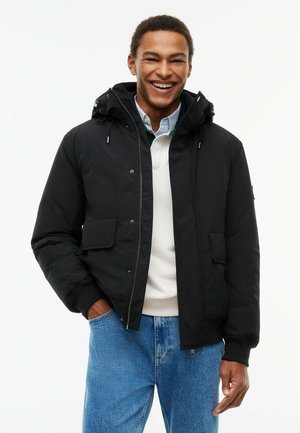 Junger Mann lächelt, trägt eine schwarze Winterjacke mit Kapuze über einem weißen Pullover und blauen Jeans, steht vor weißem Hintergrund.