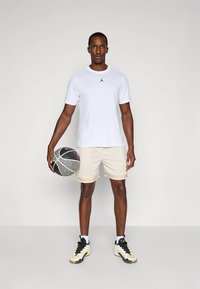 T-shirt branca de mangas curtas com logótipo preto, calções de rede bege, a segurar uma bola de basquetebol, calçando ténis atléticos pretos e brancos. Fundo neutro.