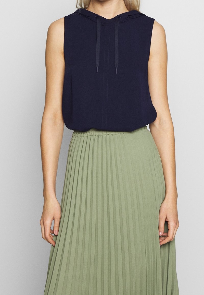 Haut sans manches bleu marine à capuche avec cordons, associé à une jupe plissée vert olive, mettant en avant des textures douces et des couleurs contrastées.