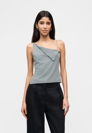 Donna con capelli lunghi e scuri che indossa una maglia grigia senza maniche asimmetrica e pantaloni neri larghi, in piedi davanti a uno sfondo bianco semplice.