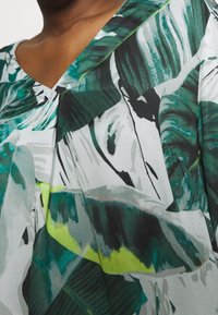 Krótki rękaw top z zielono-białym wzorem liści, z dekoltem w serek i miękką, płynną fakturą tkaniny, podkreślając jasne żółte akcenty.