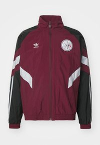 Casaco corta-vento marrom e preto com punhos elásticos, fecho zip, logótipo bordado e painéis brancos nas mangas.