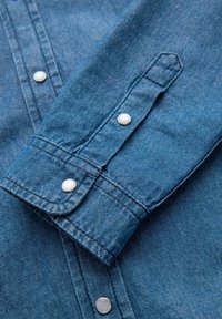 Détail d'une chemise en denim de couleur bleue, avec des boutons-pression, un poignet plié et un tissu texturé. Coutures visibles le long des bords.