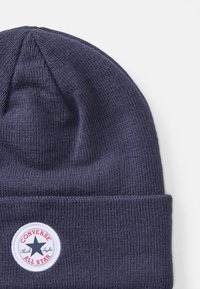 Donkerblauwe gebreide muts met een omgeslagen rand. Beschikt over een ronde patch met een sterlogo en de tekst "Converse All Star" in rood en wit.