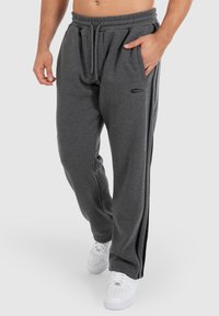Graue Sweatpants aus weichem Stoff, mit einem elastischen Bund, seitlichen Taschen und schwarzen Seitenstreifen. Getragen mit weißen Sneakern.