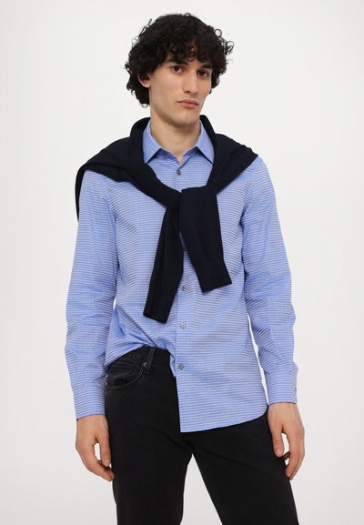 Camisa de botones azul claro con finas rayas blancas, combinada con un suéter azul marino atado alrededor de los hombros y pantalones negros.