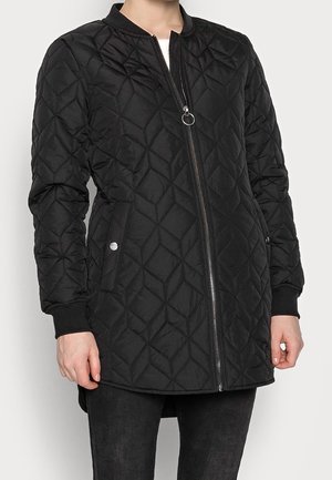 Femme portant une longue veste matelassée noire avec fermeture éclair sur le devant et poches à boutons-pression, assortie à un pantalon noir.