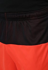 Shorts de sport noirs et orange avec une ceinture en mesh et un tissu orange vif et lisse, avec des détails de surpiqûres contrastées.