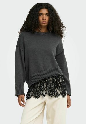Mujer con cabello largo y rizado lleva un suéter de punto gris oscuro superpuesto sobre una blusa de encaje negra y pantalones blanco hueso sobre un fondo liso.