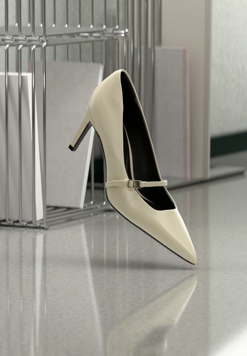 Κρεμ παπούτσι με ψηλό τακούνι και λεπτό λουράκι, τοποθετημένο σε αντανακλαστικό δάπεδο κοντά σε μεταλλικό οργανωτή αρχείων με χαρτιά.