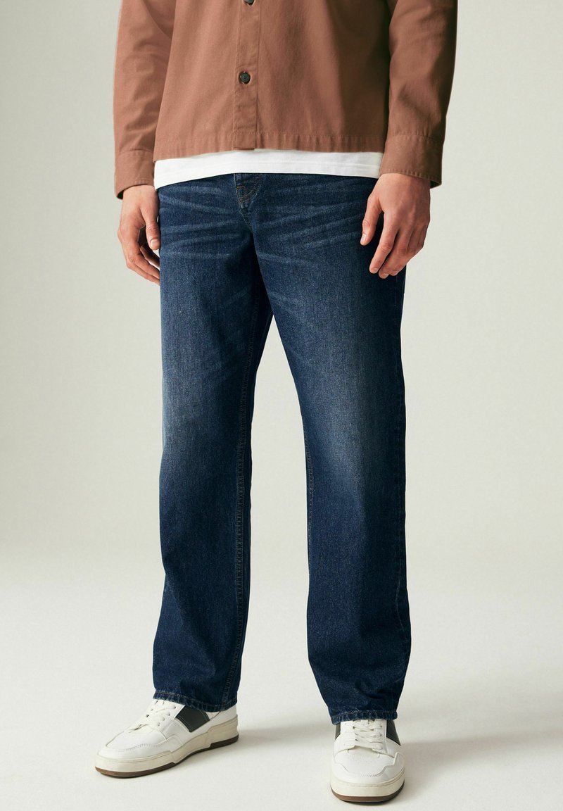 Jeans de mezclilla azul oscuro con corte recto, que presentan un ligero efecto de desgastado y detalles de costura, combinados con zapatillas blancas.