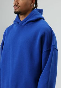 Blauwe capuchontrui van zacht stof. Heeft een losse pasvorm, doorgezakte schouders en geen zichtbare logo's of patronen.