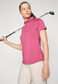 Ružičasta polo majica s ovratnikom, kratkim rukavima i gumbima; model drži golf štap, nosi svijetlosmeđe hlače.