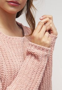 Hellrosa Pullover mit strukturiertem Strickmuster, weitem Ausschnitt und gerippten Bündchen. Der Stoff wirkt weich und plüschig.