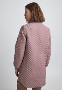 Dlouhá růžová prošívaná bunda s chevronovým vzorem, stojáčkem a žebrovanými manžetami. Design má mírně zaoblený lem.