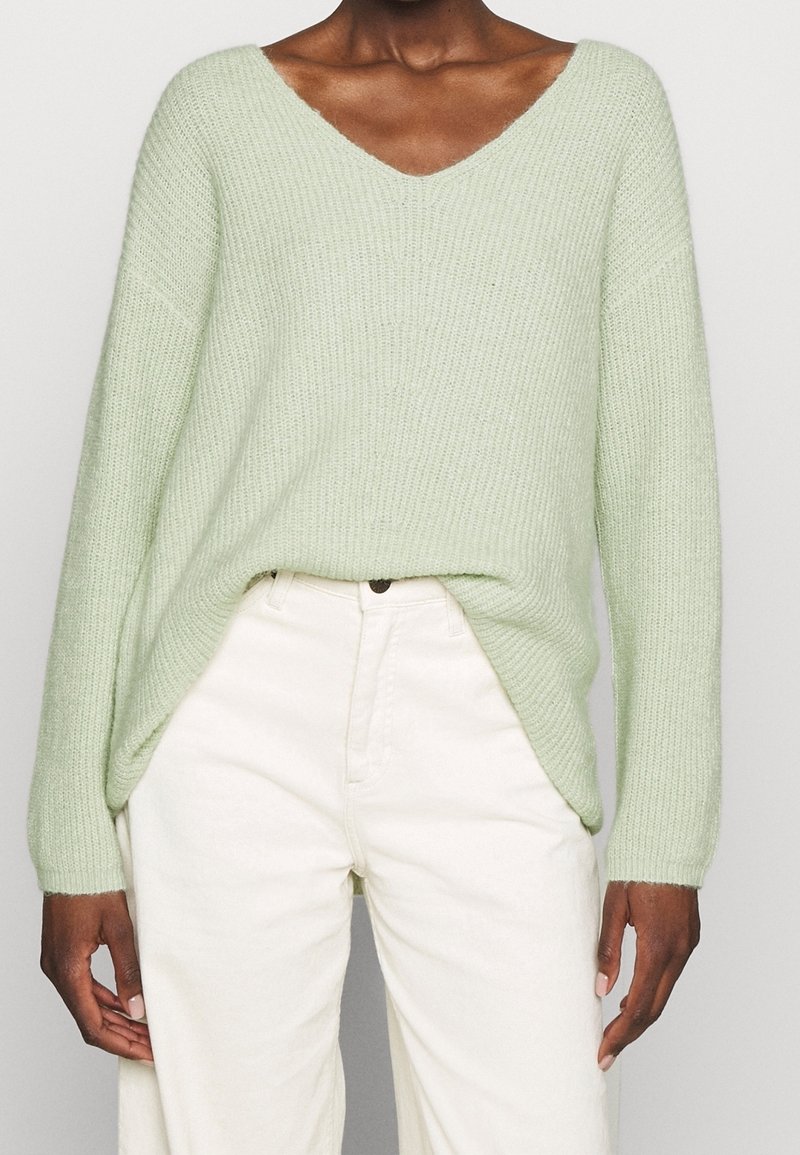Personne portant un pull en tricot vert clair à col en V, rentré dans un pantalon taille haute écru, debout devant un fond uni.