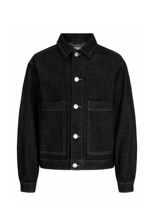 Chłopiec stoi i patrzy w bok, ubrany w czarną kurtkę dżinsową i szerokie spodnie dżinsowe na białej koszuli i beżowych butach.