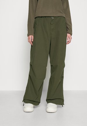 Personne portant un pantalon large vert olive avec une taille et des poignets à cordon, associé à des chaussures blanches pelucheuses à enfiler et un haut à manches longues.