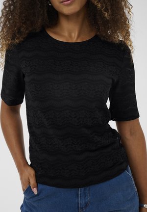 Schwarzes kurzärmeliges Oberteil mit einem strukturierten, wellenförmigen Muster. Rundhalsausschnitt und leicht lockere Passform. Kombiniert mit blauen Jeansshorts.