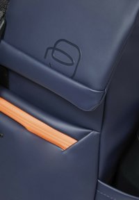 Sac à dos bleu marine avec une finition mate lisse, doté d'une poche accent orange contrastante et d'un logo discret sur le rabat avant.