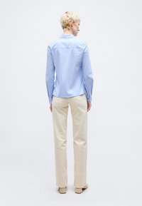 Άτομο με κοντά ξανθά μαλλιά φοράει ανοιχτό μπλε μακρυμάνικο πουκάμισο, μπεζ παντελόνι και μπεζ παπούτσια, στέκεται γυρισμένο ανάποδα σε λευκό φόντο.