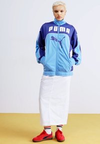 Blå og lilla treningsjakke med "PUMA"-logo, parret med et hvitt skjørt og røde sneakers med svarte og hvite stripete sokker.