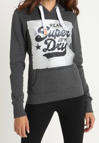 Felpa grigia scura con lacci bianchi e pannello in paillettes argento che riporta la scritta "Real Super Dry JPN", indossata con leggings neri.