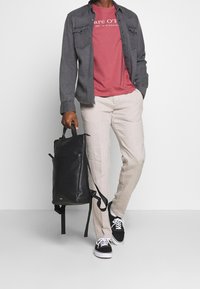 Czarny skórzany plecak z górnym uchwytem i regulowanymi paskami, noszony z szaryą koszulą zapinaną na guziki, czerwoną koszulką z grafiką i beżowymi spodniami.