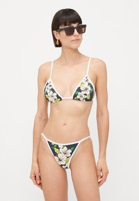 Woman wearing floral bikini with white trim, square dark sunglasses, and gold earrings against a plain white background.